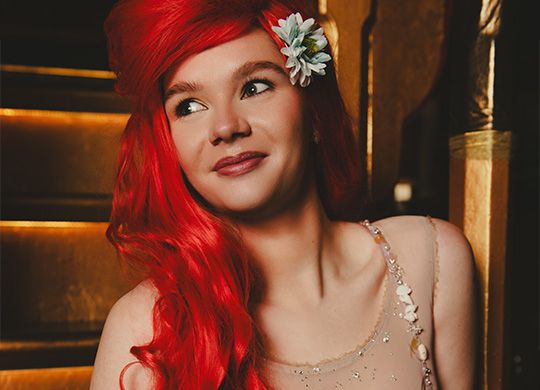 a woman with long red hair and a flower in her hair gazes thoughtfully while sitting on stairs wearing a sparkly outfit showcasing beauty and elegance for the fourth character option